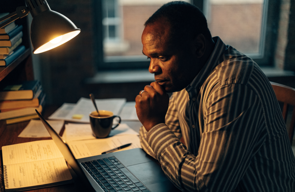 A Black man working late at his desk at home, laptop open, notebook and papers under a warm lamp