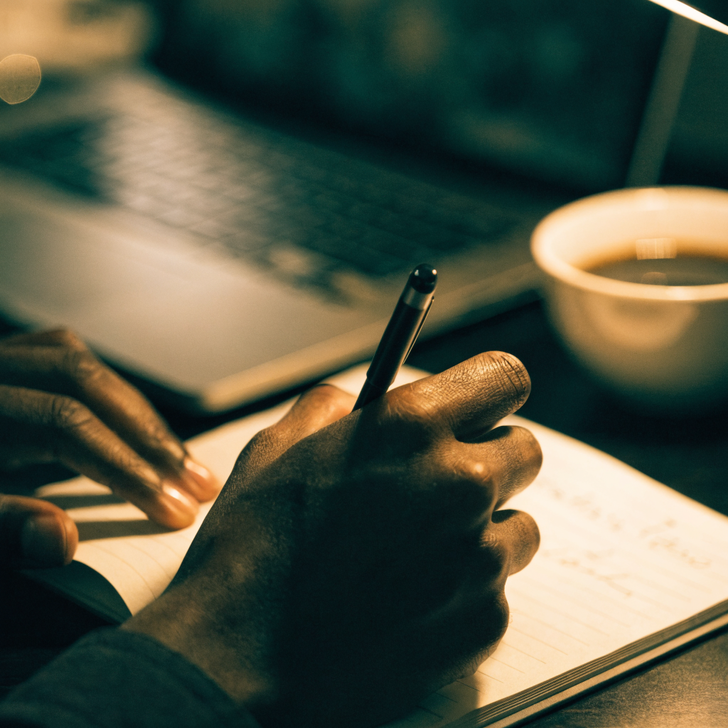 Hands writing in a notebook beside an open laptop under warm lamp light