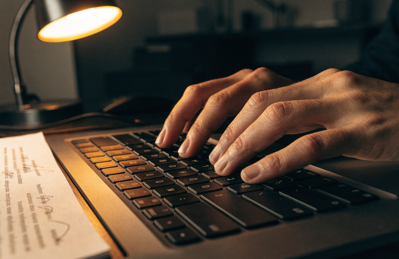 Hands typing deliberately on a laptop beside a page of handwritten notes