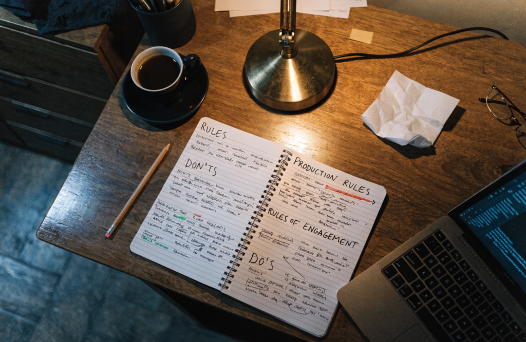 An open notebook filled with handwritten rules and headings beside a laptop and a coffee cup