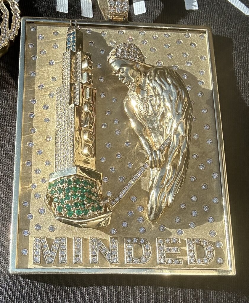 The Chicago Minded pendant in gold: a man digging, the shovel having pulled the Willis Tower up out of the ground, the blade set in green stones, with MINDED spelled across the bottom