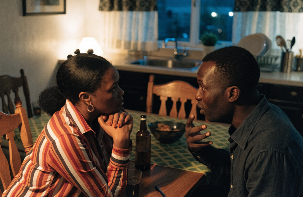 A Black couple sitting at a kitchen table mid-conversation, one listening closely, warm evening light