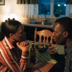 A Black couple sitting at a kitchen table mid-conversation, one listening closely, warm evening light