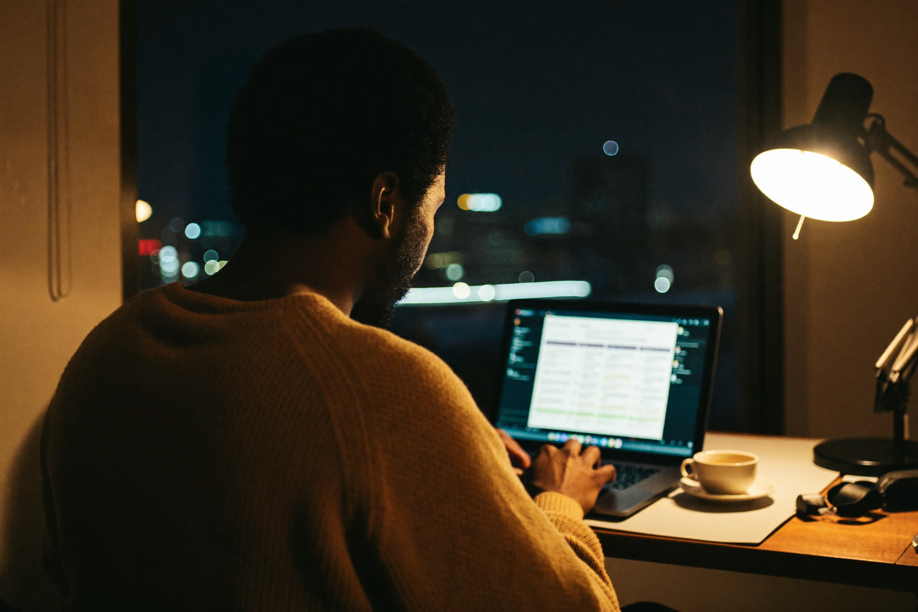 A man working late at his home desk with the city lights outside