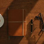 A notebook, pen and coffee on a wooden desk in afternoon light