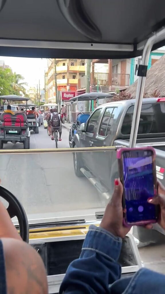 A busy main street in San Pedro, Belize seen from a golf cart, with other carts, a cyclist, a truck and people walking